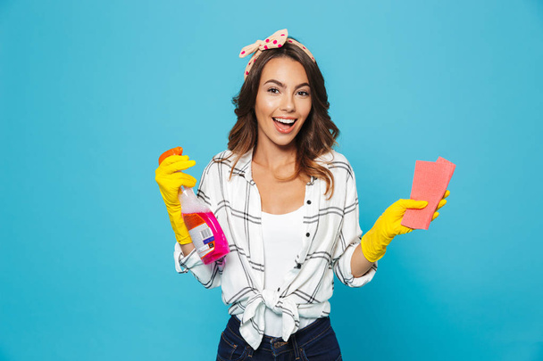 Portrait of young woman 20s housekeeping and cleaning with sponge and detergent sprayer wearing yellow rubber gloves for hands protection isolated over blue background - Photo, image