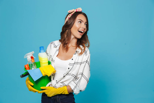 Image closeup of cheerful woman 20s wearing yellow rubber gloves for hands protection holding bucket with detergents isolated over blue background - Foto, Imagen
