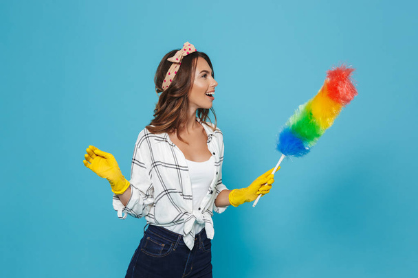 Photo of female houseworker 20s in yellow rubber gloves for hands protection holding colorful duster brush during dusting isolated over blue background - Foto, Imagen