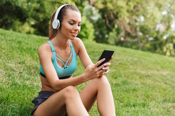 Sorrindo jovem esportista vestido sportswear em fones de ouvido descansando enquanto sentado na grama com telefone celular ao ar livre
 - Foto, Imagem