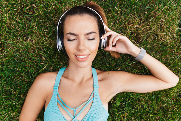 Top view of smiling young sportswoman dressed sportswear in headphones resting while laying on grass outdoors - Photo, Image