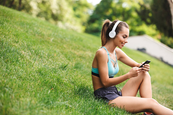Satisfied young sportswoman dressed sportswear in headphones resting while sitting on grass with mobile phone outdoors - Photo, image