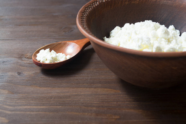 Cup of cottage cheese, wooden spoon on dark wooden background. Side view - 写真・画像