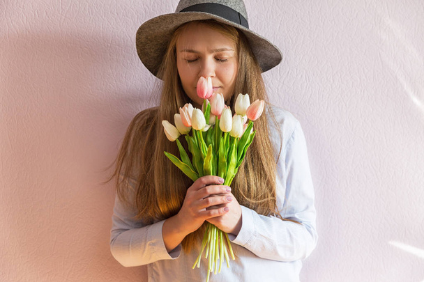 Een jong mooi meisje met blond opgeloste lange haren, een vilten hoed op haar hoofd, houdt Lentebloemen in haar handen op een zonnige dag. Dag van de vrouw. - Foto, afbeelding