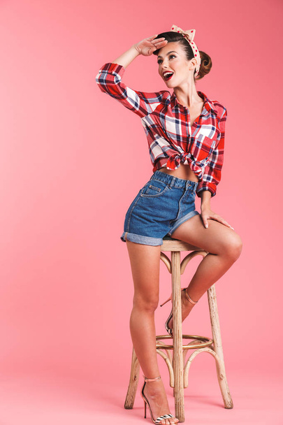 Full length portrait of a cheerful brunette pin-up girl in plaid shirt looking away while posing on a chair over pink background - Photo, image