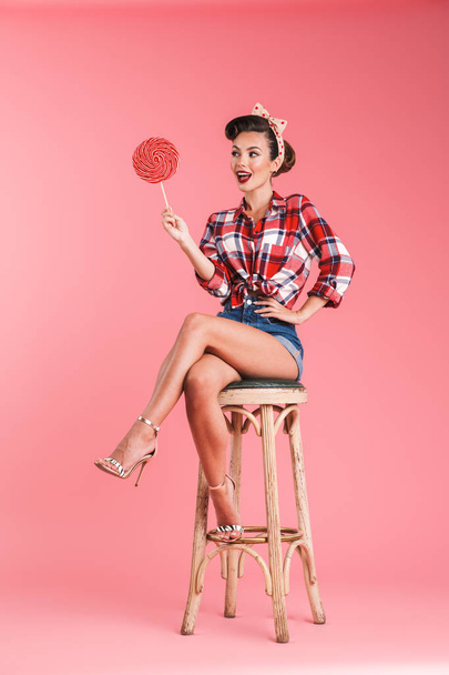 Image of gorgeous young pin-up woman sitting on stool isolated over pink background wall holding candy lollipop. - Fotoğraf, Görsel