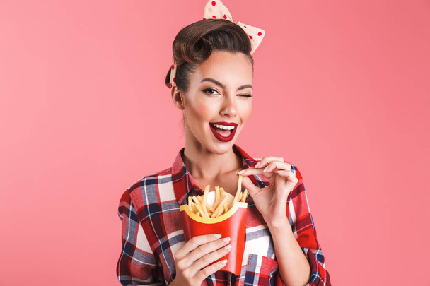 Portrait of a playful beautiful brunette pin-up woman in headband eating french fries isolated over pink background - Foto, Imagem