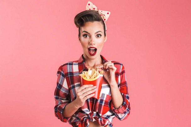 Portrait of an excited beautiful brunette pin-up woman in headband eating french fries isolated over pink background - Foto, Imagem