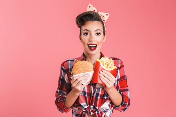 Portrait of an excited beautiful brunette pin-up woman in headband holding french fries and a burger isolated over pink background - Photo, Image