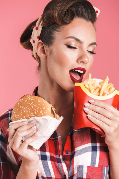 Picture of amazing young pin-up woman isolated over pink background wall holding french fries and burger. - Photo, Image