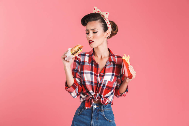 Image of sad hungry young pin-up woman isolated over pink background wall holding french fries and burger. - Photo, Image