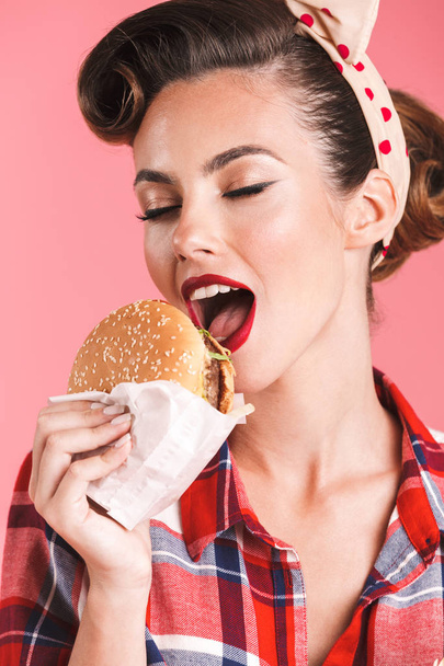 驚くほど若いピンナップ女性ハンバーガーを食べるピンクの背景の壁に分離の画像. - 写真・画像