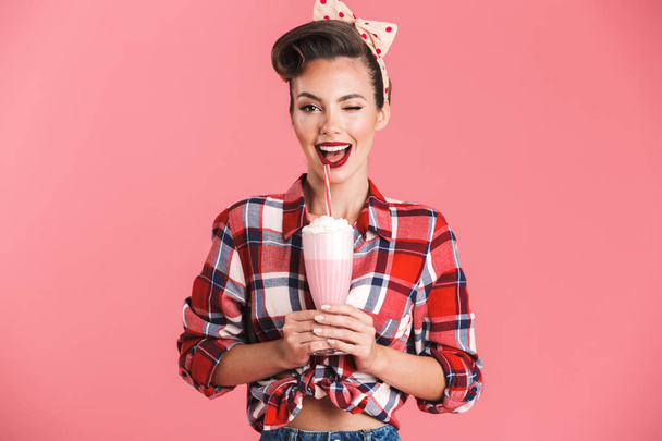 Portrait of a cheerful brunette pin-up girl in plaid shirt holding milk shake and winking over pink background - Foto, Imagem
