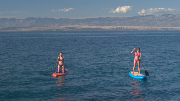 AERIAL CLOSE UP: Flying above two cheerful girlfriends in bikini standup paddle surfing in amazing blue ocean on stunning sunny summer day. High rocky mountains and arid sandy island in the background - Footage, Video