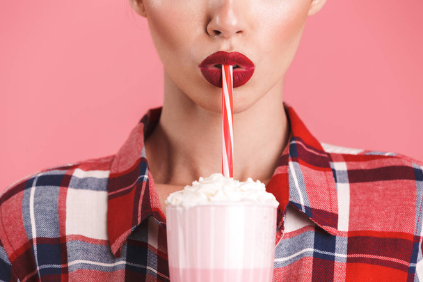 Cropped image of a cheerful brunette pin-up girl in plaid shirt drinking strawberry milk shake over pink background - Foto, Imagen