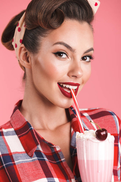 Close up portrait of a cheerful brunette pin-up girl in plaid shirt drinking milk shake over pink background - 写真・画像
