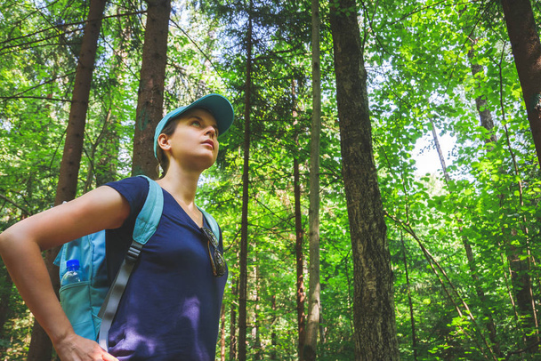 Νέοι ταξιδεύουν κορίτσι με μπλε καπάκι και σακίδιο στέκεται και κοιτώντας ψηλά σε δέντρα με πράσινο δάσος ηλιόλουστη καλοκαιρινή μέρα - Φωτογραφία, εικόνα