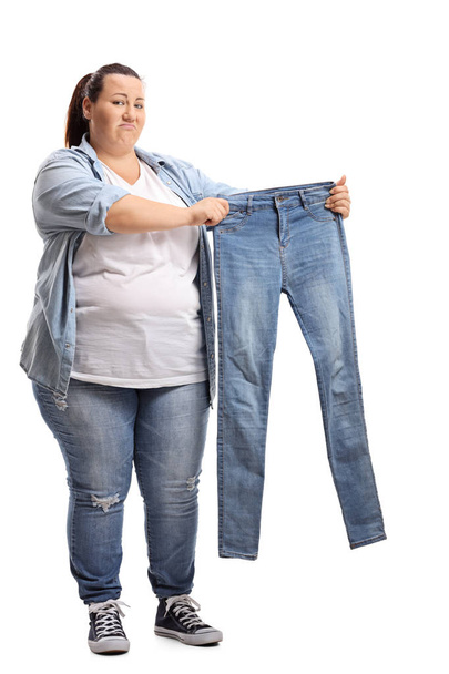 Full length portrait of an overweight woman holding a pair of small jeans isolated on white background - Foto, Imagem