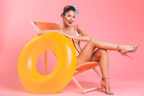 Photo of young pin-up woman isolated over pink background wall in swimwear on beach chair with inflatable ring. - Fotó, kép
