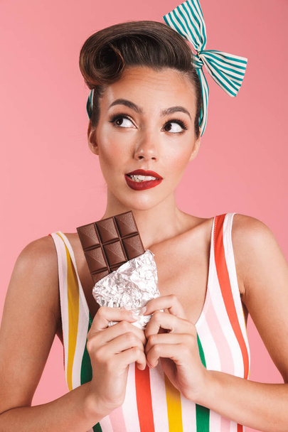Image of dreaming hungry young pin up woman isolated over pink wall background eating chocolate. - Фото, изображение