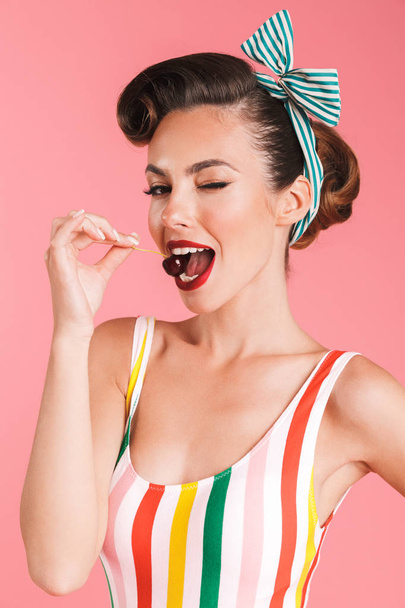 Portrait of a playful brunette pin-up girl in swimsuit eating a cherry over pink background - Фото, изображение