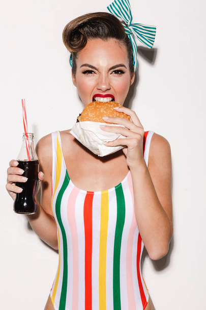 Image of beautiful pin up woman isolated over white wall background eating burger drinking soda. Looking camera. - Foto, imagen