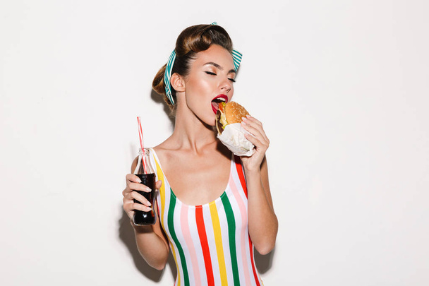 Image of amazing pin up woman isolated over white wall background eating burger drinking soda. - Photo, Image