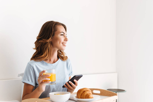 Smiling young woman holding mobile phone while having breakfast on a tray in bed - Fotografie, Obrázek