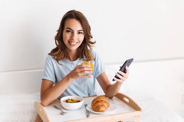 Iloinen nuori nainen käyttää matkapuhelinta, kun aamiainen tarjotin sängyssä
 - Valokuva, kuva