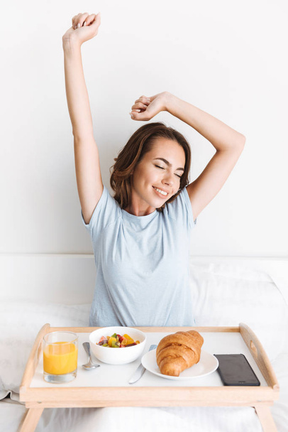 Happy young woman stretching her hands while having breakfast on a tray in bed - Fotoğraf, Görsel