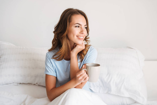 Smiling young woman holding mug while sitting in bed in the morning at home and looking away - Fotografie, Obrázek