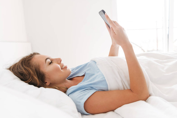 Happy young woman using mobile phone while laying in bed - Photo, image