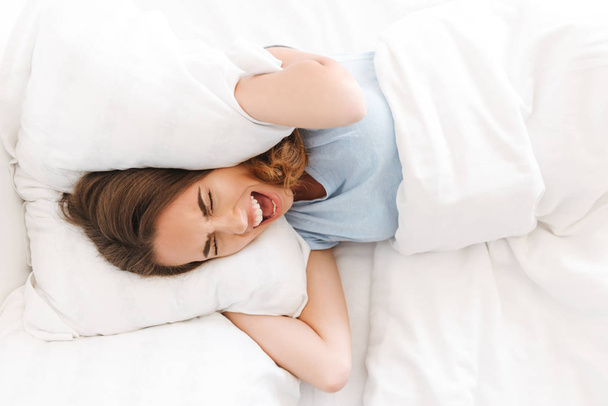 Annoyed young woman covering head with a pillow and screaming while laying in bed - Foto, immagini