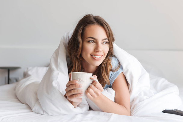 Smiling young woman holding mug while laying in bed in the morning at home - Foto, Imagen