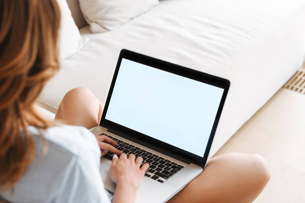 Back view of a young woman typing on blank screen laptop computer while sitting on a couch at home - Foto, Bild