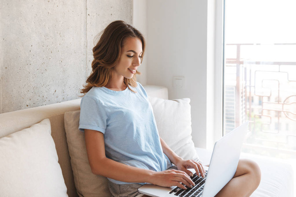 Happy young woman sitting on a couch at home with laptop computer - Foto, Bild