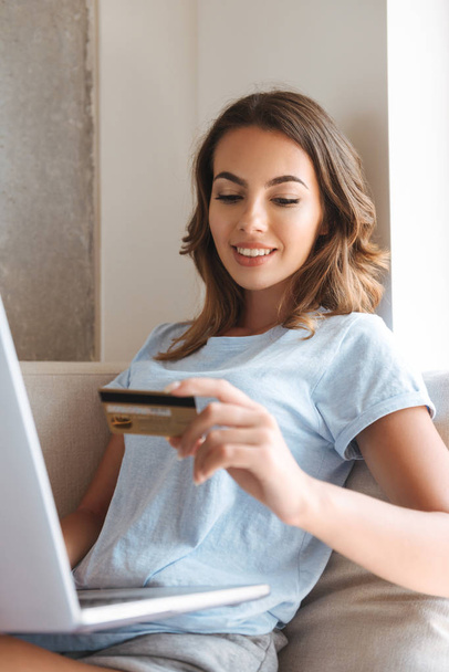 Smiling young woman holding credit card while sitting on a couch at home with laptop computer - Φωτογραφία, εικόνα