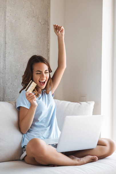 Excited young woman holding credit card while sitting on a couch at home with laptop computer - Φωτογραφία, εικόνα