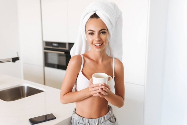 Cheerful young woman in bra and towel around her head standing on a kitchen and drinking coffee - Фото, изображение