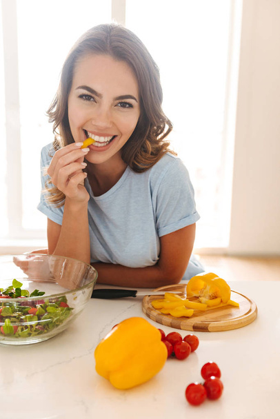 Happy young woman eating salad while sitting at the kitchen table - Foto, afbeelding