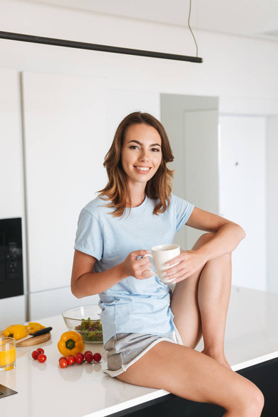 Delighted young woman drinking coffee while sitting at the kitchen at home in the morning - Foto, Imagen