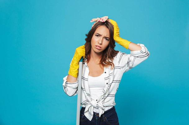 Portrait of a tired housewife standing with a broom looking at camera isolated over blue background - Fotografie, Obrázek