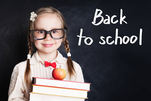 Glimlachend kind meisje in school uniform houden van boeken en apple tegen krijt bestuur achtergrond. Terug naar school concept - Foto, afbeelding