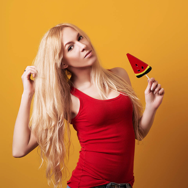 Young sexy blonde fashion girl with long hair in red top, holding lollipop over yellow background. Copy space. Sexy young woman with candy. Female fashion. Seasonal shopping, sales concept - Photo, Image