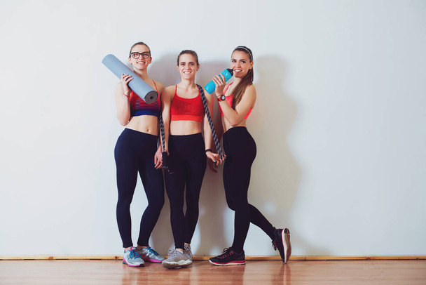 Young sporty women after training in fitness studio. Fitness, sport and healthy lifestyle concept. Group of females in sportswear standing together in front of white wall. Girls fitness exercise - Photo, Image