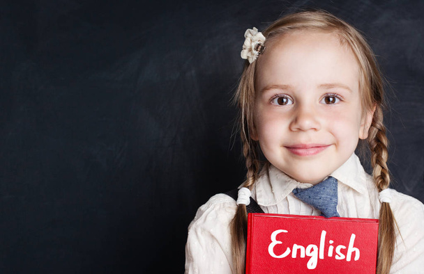 Τα παιδιά μαθαίνουν αγγλικά έννοια. Closeup πορτρέτο της χαριτωμένο παιδί κορίτσι με Ελληνικά βιβλίο για μαυροπίνακα σε κέντρο ξένων γλωσσών - Φωτογραφία, εικόνα