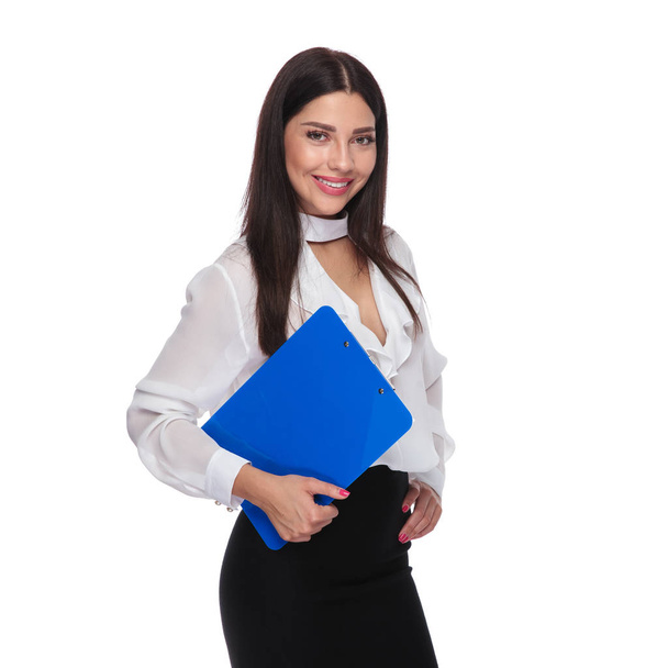 Porträt einer jungen brünetten Geschäftsfrau mit einem blauen Klemmbrett, während sie auf weißem Hintergrund steht und ihre Hüfte hält - Foto, Bild