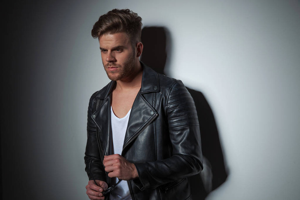 portrait of sexy man wearing leather jacket looks down to side while leaning against a grey wall, holding sunglasses - Foto, Imagem