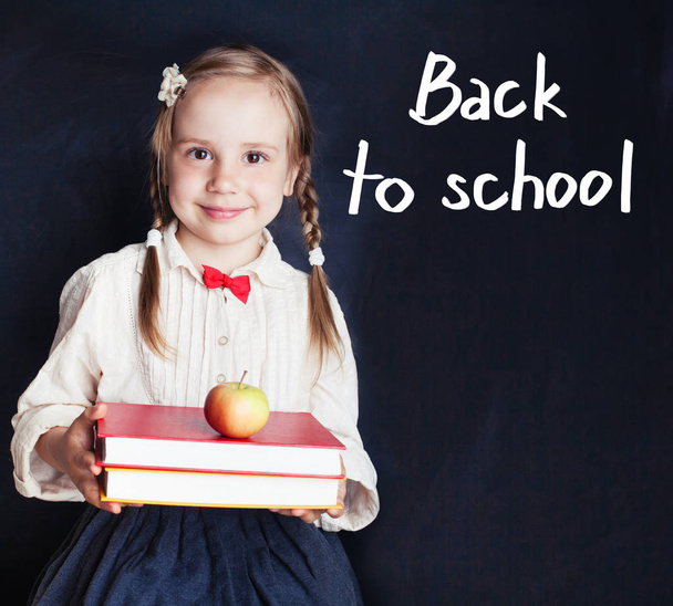 Kleine Schülerin in Schuluniform lächelt und hält einen Stapel Bücher und einen Apfel auf Schultafel-Hintergrund mit Schulhandschrift - Foto, Bild