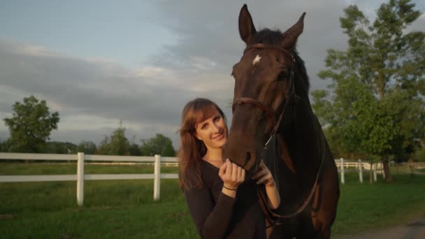 Lassú mozgás közelről: Gyönyörű női lovas alsószoknya és csók a lenyűgöző az sunny reggeli előtt a vihar erős sötét barna ló. Csinos lány élvezi a nyári lovaglás vakáció a természetben - Felvétel, videó